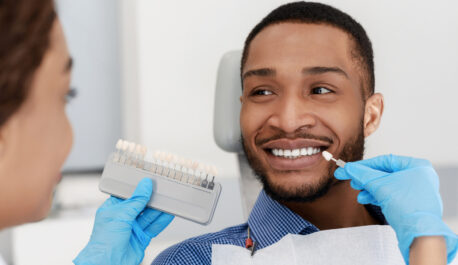 Female dentist choosing filling shade for smiling black guy, using tooth scale sample, close up